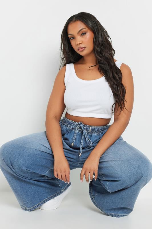 Woman wearing a white tank top and blue jeans sitting on a white background