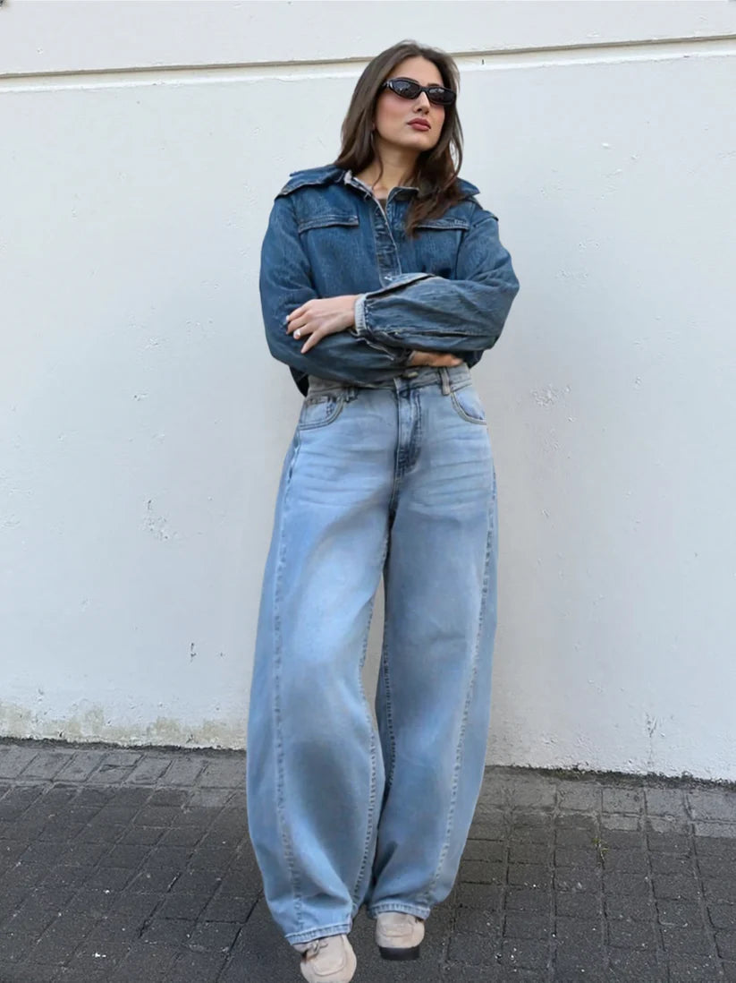 Woman wearing a denim jacket and jeans standing against a white wall.