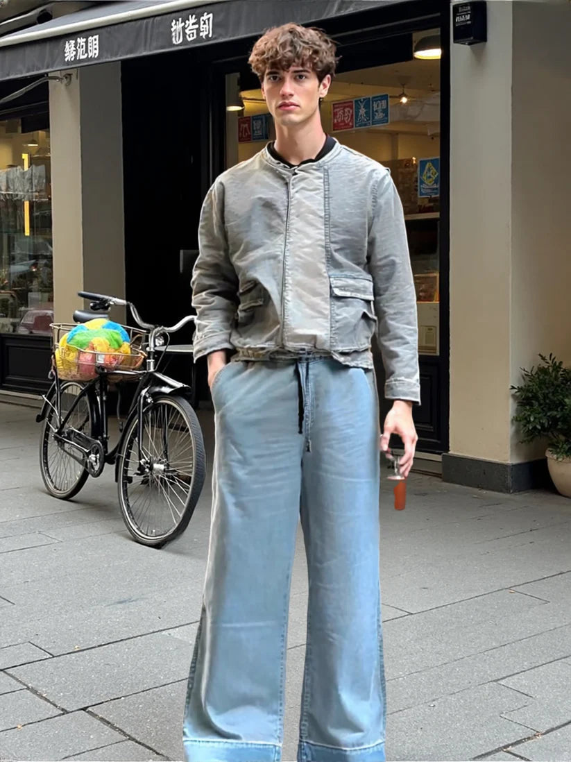 Person wearing a gray jacket and light blue jeans standing on a street with a bicycle and store in the background.